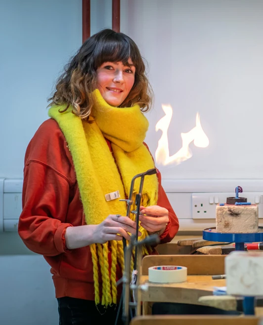 A female jewellery student in a red sweater and yellow scarf using a torch to heat metal, creating a visible flame in the workshop. A female jewellery student in a red sweater and yellow scarf using a torch to heat metal, creating a visible flame in the workshop.